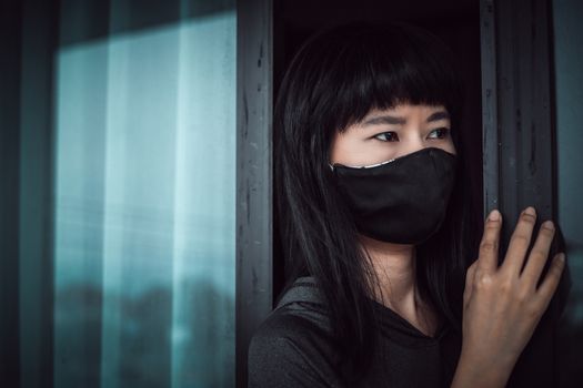 Asian woman wearing a black face mask, standing absent-minded at the door and staying home for self-quarantine and social distancing in coronavirus or Covid-2019 outbreak situation concept