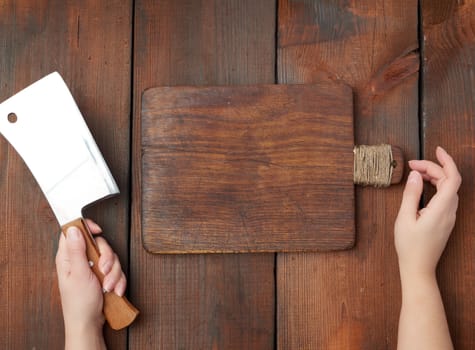 very old empty wooden rectangular cutting board and hands hold a large sharp meat knife, top view, brown wooden background 