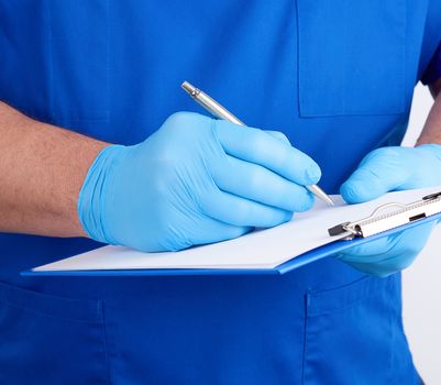 doctor in blue uniform and latex gloves holds a blue holder for sheets of paper and writes with his right hand, white background