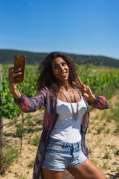 Pretty young Brazilian woman taking a selfie photo with a smartphone outdoors in strong light. Girl smiling expressing energy in good day. Lovely curly woman with good vibes