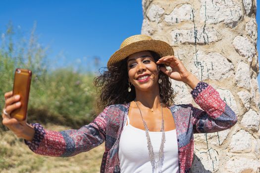 Pretty young Brazilian woman taking a selfie photo with a smartphone outdoors in strong light. Girl smiling expressing energy in good day. Lovely curly woman in stylish hat