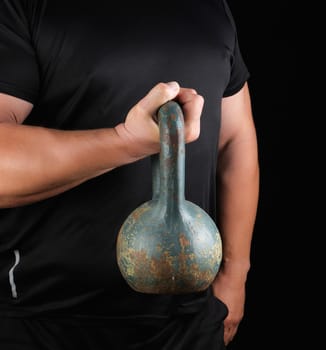 adult strong athlete in black clothes holding an iron kettlebell on his outstretched arms, black background