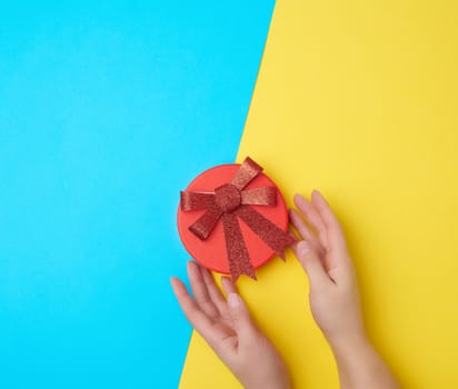 two female hands hold a closed paper cardboard box with a red bow on a yellow-blue background, concept of surprise