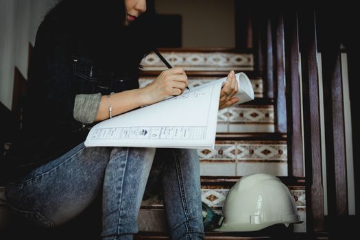 Asian woman looking architectural plan, sitting on the stairs and working at home with safety helmet for building inspection concept