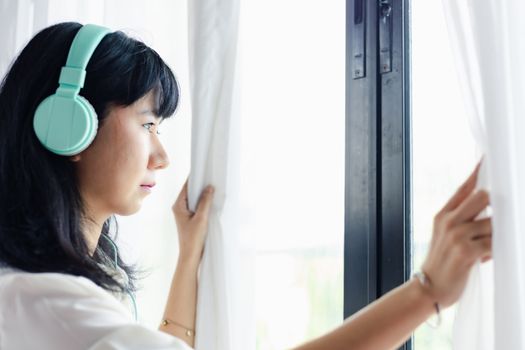 Asian woman staying home, wearing headphones and standing absent-minded at the door for self-quarantine and social distancing in coronavirus or Covid-2019 outbreak situation concept