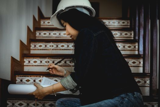 Asian woman looking architectural plan, sitting on the stairs and working at home with safety helmet for building inspection concept