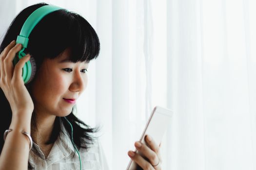 Asian smiling woman wearing headphones, listening music and using smartphone for entertainment and leisure at home concept