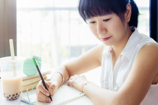Asian woman writing on a notebook with a plastic cup of bubble tea against blurred background for staying and working at home concept