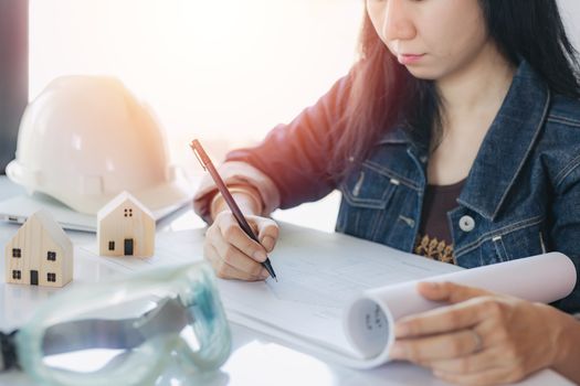 Asian woman checking architectural plan with personal safety equipment and wooden house model on white office desk for building inspection concept