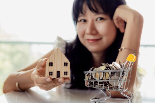 Selective focus of asian woman's hand holding a wooden house model with full of coins in shopping cart for planning, savings and property investment concept