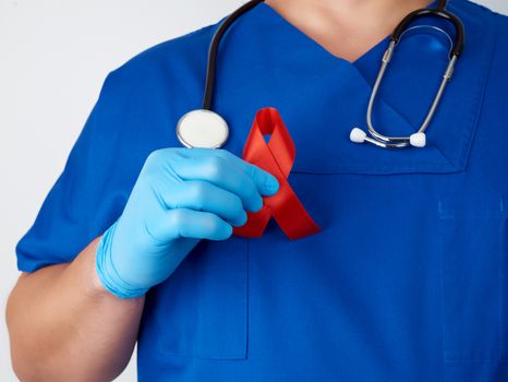 doctor in blue uniform and sterile latex gloves holds a red ribbon - a symbol of the fight against disease AIDS and vasculitis