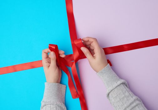 two female hands tie a bow with a satin red ribbon on a gift box, top view