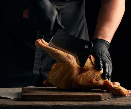 chef in black uniform and latex gloves chopping raw chicken into pieces on a brown wooden board, dark background, cooking process