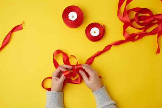 two female hands tie a bow with a red ribbon and reels with a satin ribbon on a yellow background, top view