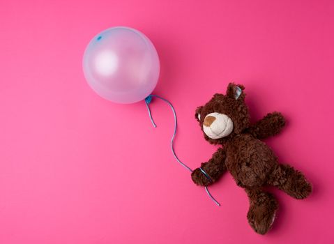 little brown teddy bear holding a blue inflated balloon on a rope on a pink background