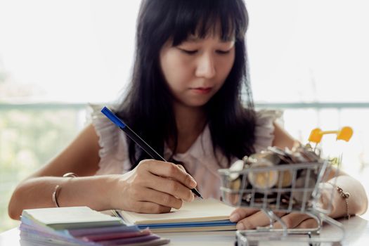 Selective focus of Asian woman's hand writing on a notebook with blurred money and coins in a shopping cart for business and financial planning concept