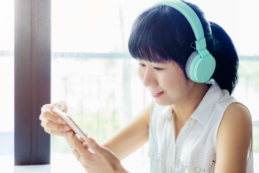 Asian smiling woman wearing headphones, listening music and using smartphone with blurred background for entertainment, relaxation and stay at home concept
