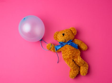 little brown teddy bear holding a blue inflated balloon on a rope on a pink background
