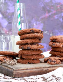 stack of round chocolate chip cookies on a brown wooden board, behind a bottle of milk