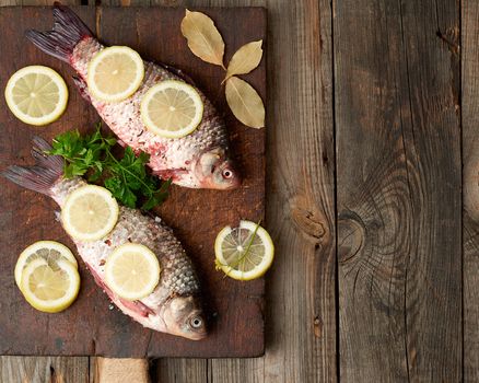 fresh crucian fish sprinkled with spices and lemon slices and lies on a brown wooden cutting board, wooden table from boards, top view