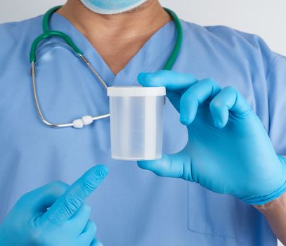 doctor in blue uniform and latex gloves is holding an empty plastic container for taking urine samples, close up