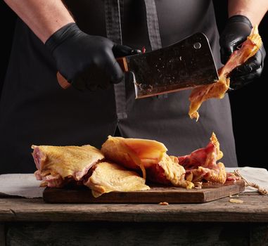 chef in black uniform and latex gloves chopping raw chicken into pieces on a brown wooden board, dark background, cooking process