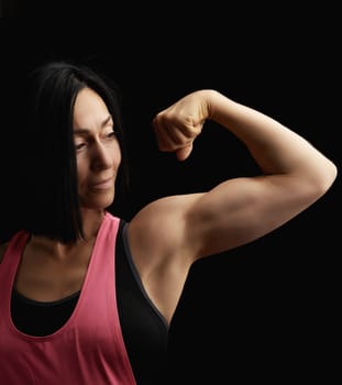 beautiful athletic girl raised and bent her arm demonstrating her biceps, athlete is standing on a dark background, wearing a black top 