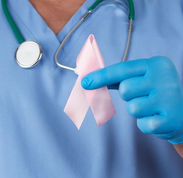 doctor in blue uniform and sterile latex gloves holds a pink ribbon a symbol of the fight against breast cancer in October every 20