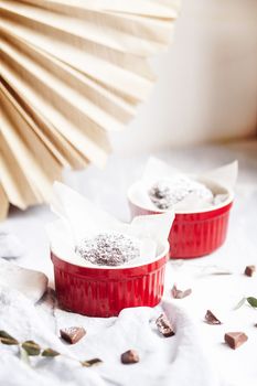 Chocolate muffins in red cups. Small glazed ceramic ramekin with brown cakes on a gray and white background
