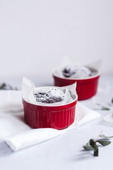 Chocolate muffins in red cups. Small glazed ceramic ramekin with brown cakes on a gray and white background