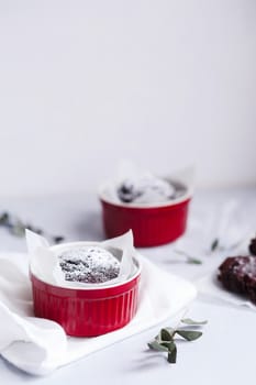 Chocolate muffins in red cups. Small glazed ceramic ramekin with brown cakes on a gray and white background