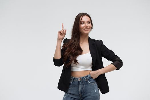Smiling amazed young brunette in black turtleneck having idea and pointing forefinger up while standing against white background