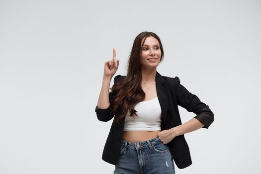 Smiling amazed young brunette in black turtleneck having idea and pointing forefinger up while standing against white background