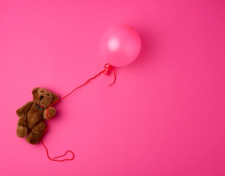 little brown teddy bear holding a pink inflated balloon on a rope on a pink background