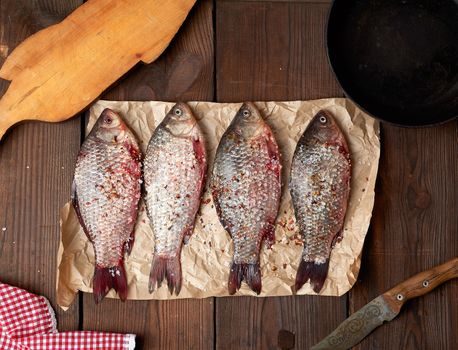 fresh crucian fish sprinkled with spices lies on brown crumpled paper, wooden table from boards, top view
