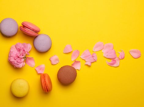 multi-colored macarons with cream and a pink rose bud with scattered petals on a yellow  background, top view, flat lay, copy space 