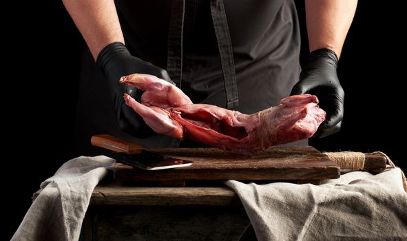 chef in black latex gloves holds a whole rabbit carcass on a brown cutting board, process of cooking meat