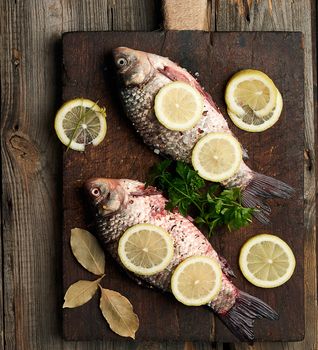 fresh crucian fish sprinkled with spices and lemon slices and lies on a brown wooden cutting board, wooden table from boards, top view