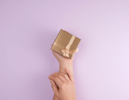 women's hands hold a closed box with a bow on a purple background, top view