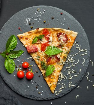 delicious triangular slice of pizza with smoked sausages, mushrooms, tomatoes, cheese and basil leaves on a black graphite board, top view