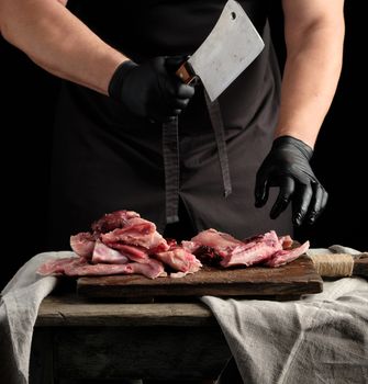 chef in black latex gloves holds a big knife and cuts into pieces raw rabbit meat on a brown wooden cutting board, cooking on a dark background