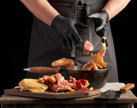chef in black uniform and latex gloves chopping throws sliced chicken into a black cast-iron frying pan with a wooden handle, meat flies into a container, dark background