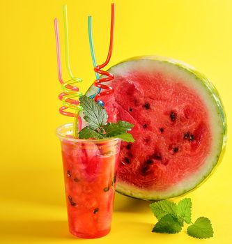 smoothie from ripe red watermelon in a plastic cup, lot of colorful tubes and a sprig of mint on a yellow background