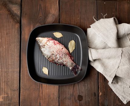 fresh crucian fish sprinkled with spices and lies in a black square pan, brown wooden table from boards, top view, copy space
