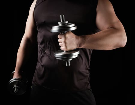 man in black clothes holds steel dumbbells in his hands, his muscles are tense, low key, sports background