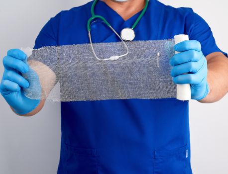doctor in blue uniform and latex gloves holds a roll of white bandage for dressing gauze wounds, white background
