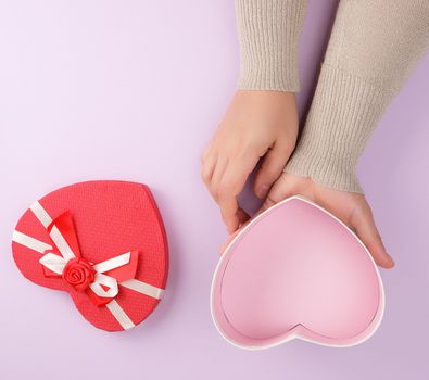 two hands hold a paper open red box in the shape of a heart on a purple background, festive backdrop, top view
