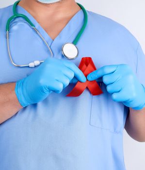 doctor in blue uniform and sterile latex gloves holds a red ribbon-symbol of the fight against disease AIDS and vasculitis, close up