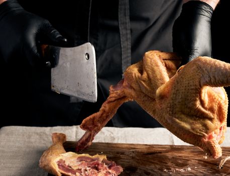 chef in black uniform and latex gloves chopping raw chicken into pieces on a brown wooden board, dark background, cooking process