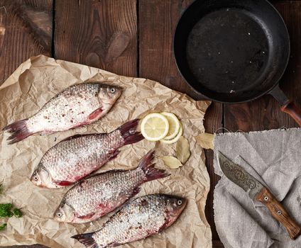 fresh crucian fish sprinkled with spices lies on brown crumpled paper, wooden table from boards, top view